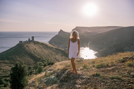 A blonde woman stands on a hill overlooking the ocean. She is wearing a white dress and she is enjoying the view