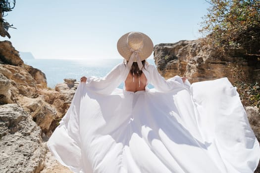 A woman in a white dress is standing on a rocky beach with her hat on. The scene is serene and peaceful, with the woman enjoying the view of the water.
