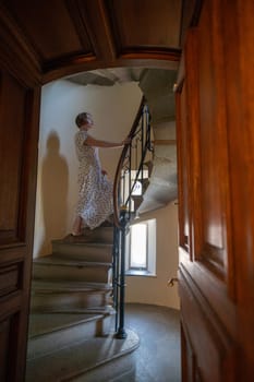 A woman is standing on a spiral staircase. The staircase is made of wood and has a metal railing. The woman is wearing a white dress and is looking up at the top of the staircase