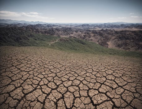 Beneath a clear blue sky, the parched ground displays extensive cracks, revealing the effects of drought. Rugged mountains extend into the horizon, creating a stark contrast against the dry terrain.