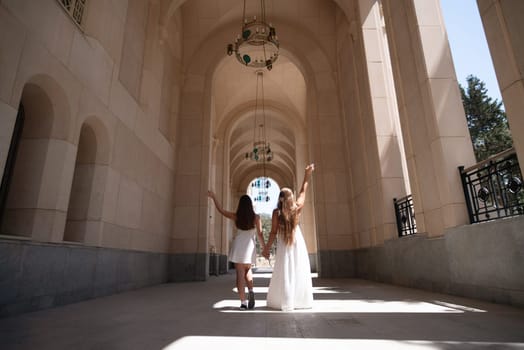 Two women in white dresses stand in an arched corridor, arms raised, facing away from the camera. Sunlight streams through the arches.