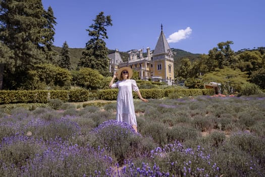 A woman stands in a field of lavender flowers. The field is surrounded by a stone wall and a large house. The woman is wearing a white dress and a straw hat. The scene is peaceful and serene