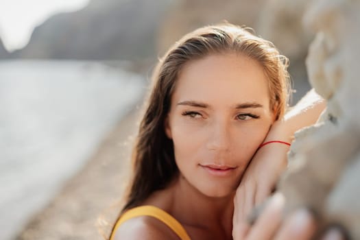 A woman with long hair is leaning against a rock on a beach. She is wearing a yellow top and a red bracelet