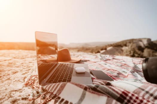 A laptop, a smartphone and a hat are arranged on a plaid blanket, overlooking a scenic landscape.