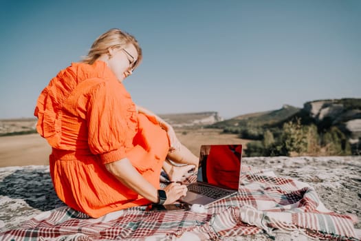 A woman in a red dress sits on a blanket on a cliff, using a laptop. The bright orange dress contrasts with the blue sky and green landscape.