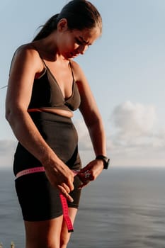 A woman is holding a pink tape measure around her waist. She is wearing black pants and a black top