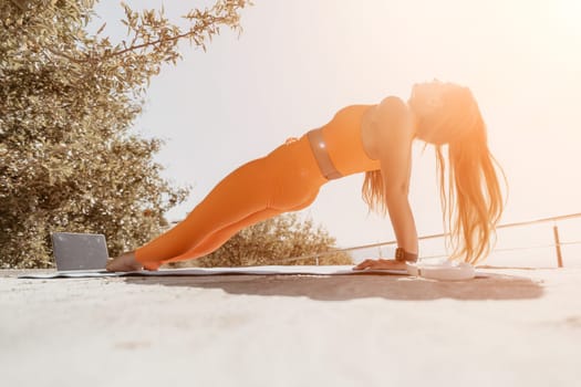 A woman in orange workout clothes performs a plank pose on a yoga mat outdoors with a laptop in the background and a tree to the left.