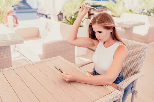 Woman sits in outdoor cafe at wooden table, holds smartphone. Bright sunny day provides natural lighting. Cafe offers relaxed atmosphere for customers to enjoy refreshments