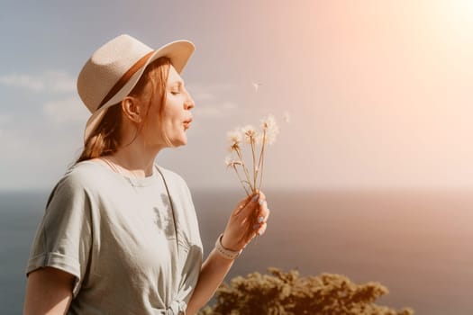 A young woman with red hair, wearing a hat, blows on a bunch of dandelions, while standing against a bright, sunny sky.