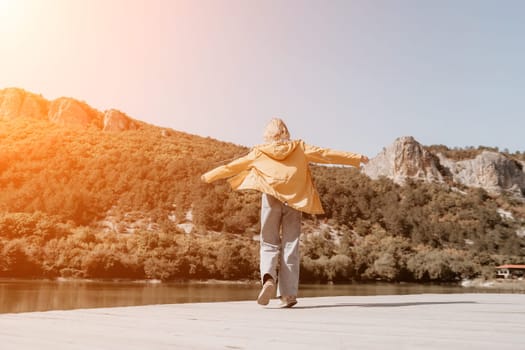A woman in a yellow jacket spins with arms outstretched, facing away from the camera, with a river and mountains in the background.