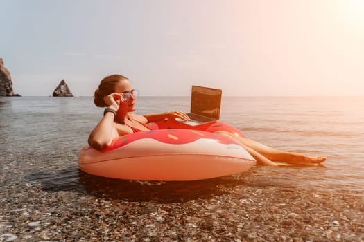 Woman freelancer works on laptop swimming in sea on pink inflatable ring. Pretty lady typing on computer while floating in the sea on inflatable donut at sunset. Freelance, remote work on vacation