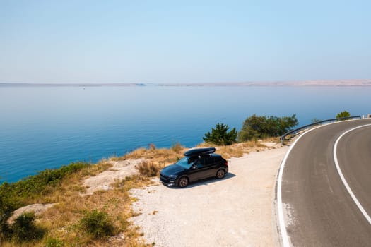 Car with a roof box, parked beside a narrow, winding road on a green cliff, with the azure sea