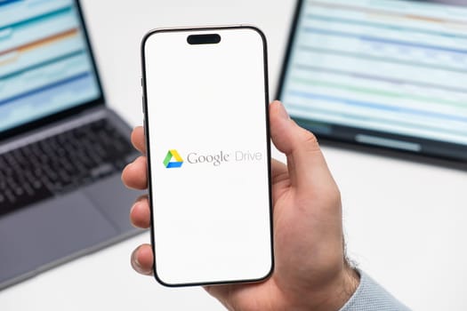 PRAGUE, CZECHIA - JULY, 2024: Google Drive logo on the screen of smartphone in mans hand and laptop on the background