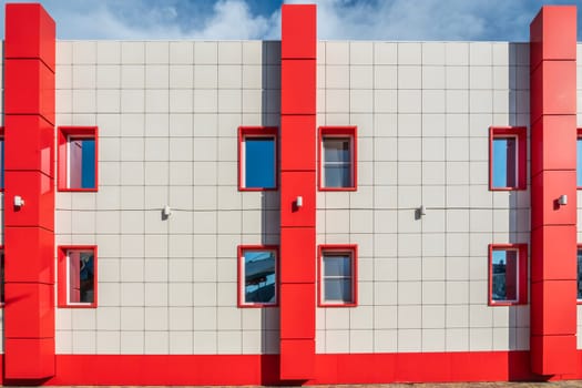 A contemporary building showcases a striking design with red and gray walls. Bright sunlight illuminates the structure, highlighting the vibrant color contrast and window placements.