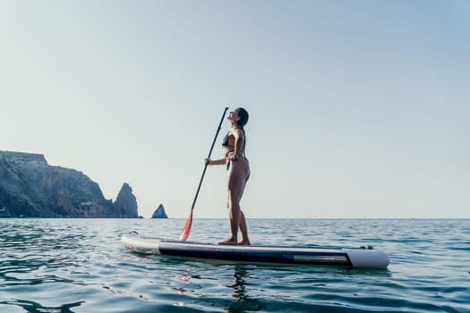 A woman stands on a paddleboard, holding a paddle, with a cliff face in the background. She is paddling out to sea.