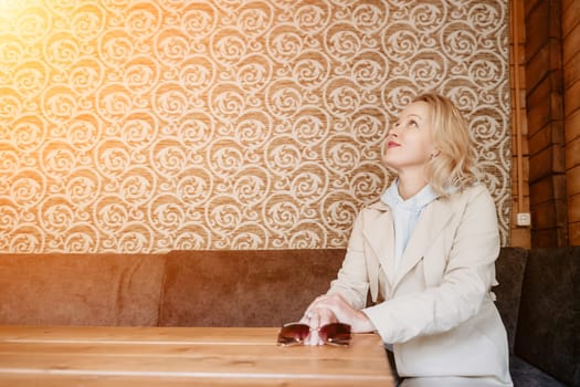 A woman with blonde hair, wearing a beige trench coat and a blue hoodie, is sitting at a table in a cafe, looking upwards thoughtfully. The background features patterned wallpaper and a wooden wall.