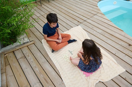 Two kids play with word tiles on a blanket by the pool. The setting evokes summer fun, relaxation, and youthful creativity.