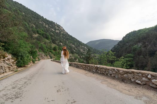 A woman is walking down a road in a mountainous area. The road is narrow and winding, and the woman is wearing a white dress. The scene has a peaceful and serene mood, with the woman's dress