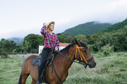 A woman is riding a horse in a field. The woman is wearing a red and white plaid shirt and a straw hat. The horse is brown and is standing in a grassy field. The scene is peaceful and serene