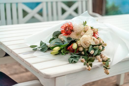 A bouquet of flowers is sitting on a table by the ocean. The flowers are white and orange, and they are arranged in a vase. The table is surrounded by chairs