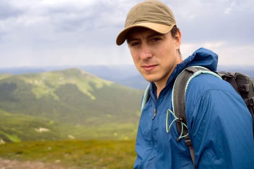 Portrait of a young traveler in tourism gear against the backdrop of beautiful mountains and hills on a sunny day. A man travels in the wild, looks into the frame, closeup view.