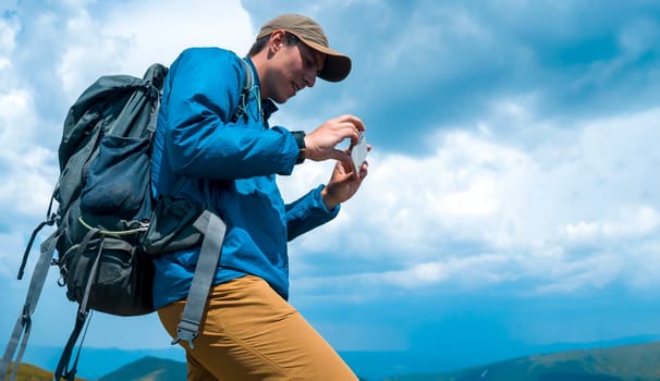 A young man is hiking, trekking in the mountains with a backpack on a sunny day, goes on adventures with travel equipment. A tourist takes a photo on the phone of beautiful landscapes.