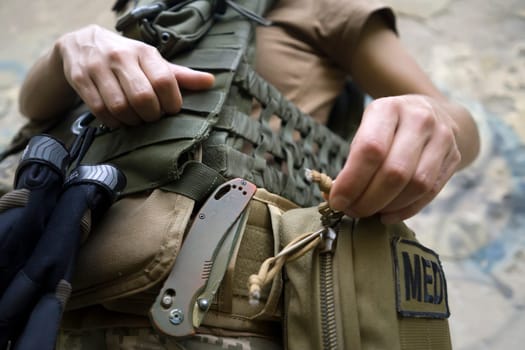 A doctor in a bulletproof vest, in camouflage, opens a first aid kit, a woman is engaged in tactical medicine and saves lives on the front line, a close-up hand.
