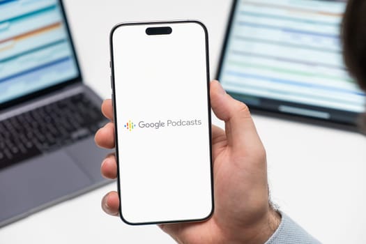 PRAGUE, CZECHIA - JULY, 2024: Google Podcasts logo on the screen of smartphone in mans hand and laptop on the background