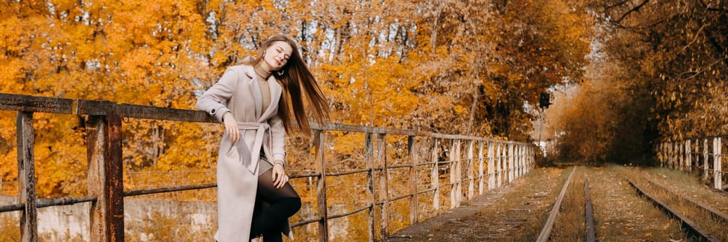 A beautiful long-haired woman walks through the autumn streets. Railway, autumn leaves, a woman in a light coat.