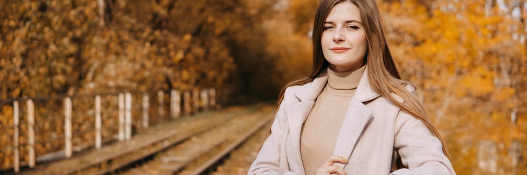 A beautiful long-haired woman walks through the autumn streets. Railway, autumn leaves, a woman in a light coat.