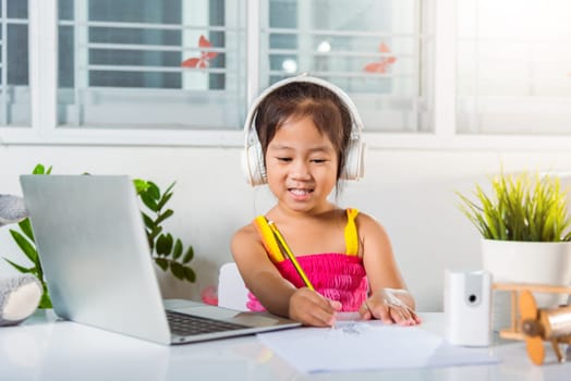 Little kid preschool wear headphone sit at desk use laptop computer and communicates on Internet online video call with teacher, Asian child girl study video conference distant education at home