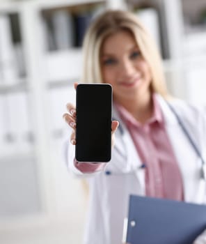 Beautiful female blonde doctor holding smartphone in hand at office woman discussing disease and giving an online consultation to a remote training mobile advertising app