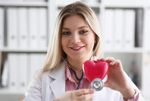 Beautiful smiling blond female doctor hold in arms red toy heart closeup. Cardio therapeutist student education CPR 911 life save physician make cardiac physical pulse rate measure arrhythmia