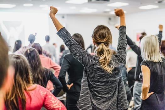 Life coaching symposium. Speaker giving interactive motivational speech at business workshop. Rear view of unrecognizable participants feeling empowered and motivated, hands raised high in air.