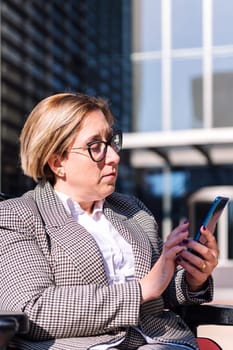portrait of a businesswoman using wheelchair typing a message on mobile phone in front of an office building in the financial district, concept of diversity and technology of communication