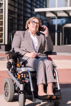 smiling businesswoman using wheelchair talking by mobile phone in front of an office building in the financial district, concept of diversity and technology of communication