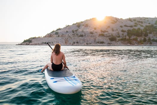 A woman enjoys paddle boarding on calm waters at sunset near a scenic coastal landscape
