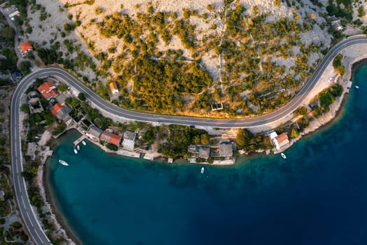 Aerial view of a coastal village with winding road and clear blue water in the afternoon sun.