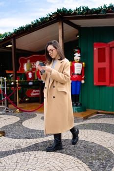 Woman in glasses using mobile phone at day in the city. Happy woman using smartphone dressed stylish trench coat looking device screen. Vertical photo full length