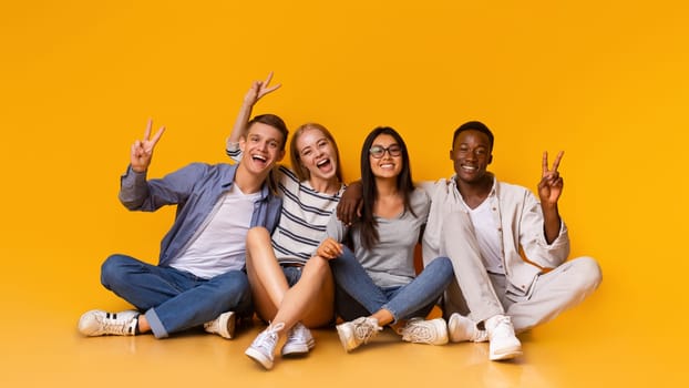 Multiracial happy students sitting on floor, embracing and having fun over yellow studio background, students lifestyle