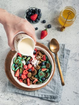 Trendy food - pancake cereal. Milk pouring on heap of multicolor mini cereal pancakes in bowl. Tiny cereal pancakes with berries in craft plate over gray cement background. Vertical. Top view,flat lay