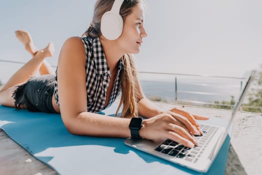 A young woman is lying on a blue mat outside with headphones on, working on a laptop. She is wearing a checkered shirt, denim shorts, and a watch.