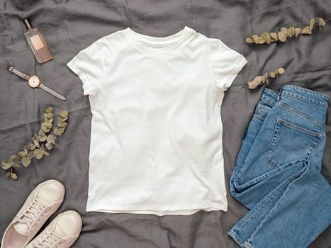 Fashionable look with white empty t-shirt, jeans and white sneakers. Top view of white blank t-shirt with short sleeves over gray bed linen. Mock up for t-shirt print design.
