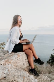 Digital nomad, woman in the hat, a business woman with a laptop sits on the rocks by the sea during sunset, makes a business transaction online from a distance. Freelance, remote work on vacation.