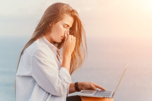 Digital nomad, woman in the hat, a business woman with a laptop sits on the rocks by the sea during sunset, makes a business transaction online from a distance. Freelance, remote work on vacation.
