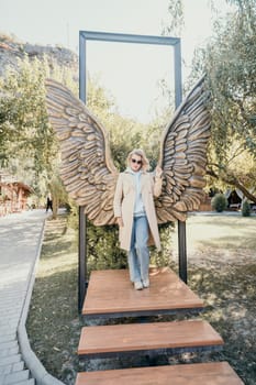 A young woman wearing sunglasses and a beige trench coat poses in front of a large angel wings sculpture in a park.