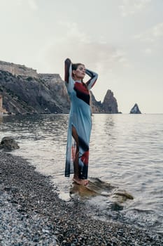 A woman in a long, colorful dress stands on a rock by the ocean, gazing at the horizon.