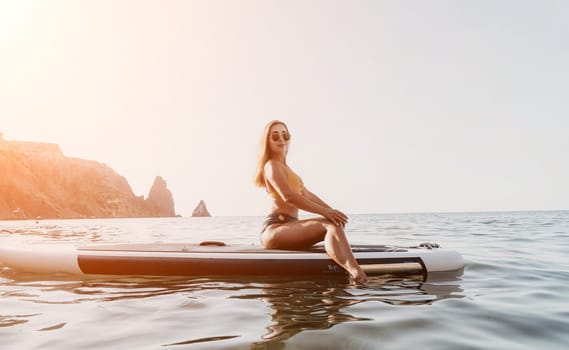 A woman in a bikini sits on a paddleboard in the ocean, looking back at the camera. The water is calm and the sky is clear.