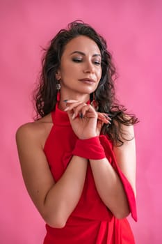 A woman in a red dress is standing in front of a pink background. She is wearing a red scarf and earrings