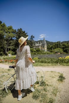 A woman in a white dress is standing next to a bicycle. She is wearing a straw hat and is looking at the camera. The scene is set in a garden, with a house in the background